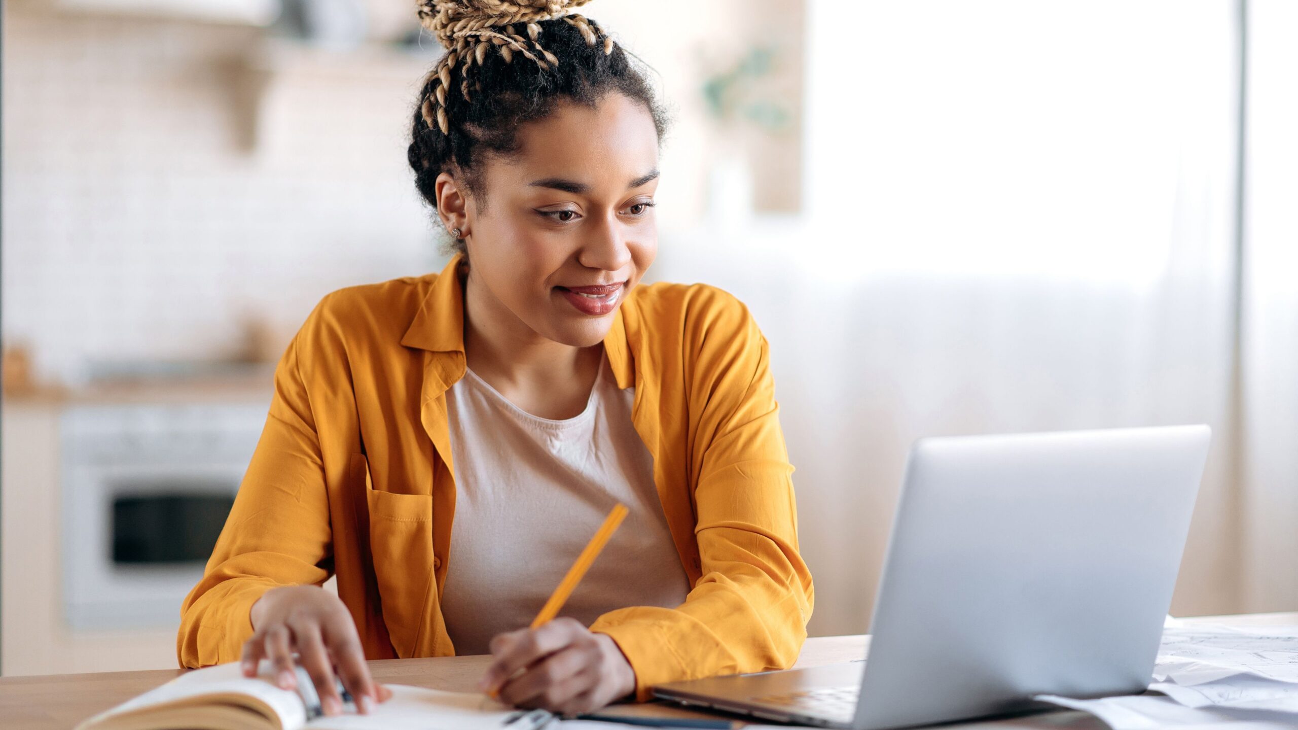 Focused African American female student with afro dreadlocks, studying remotely from home, using a laptop, taking notes on notepad during online lesson, e-learning concept, smiling