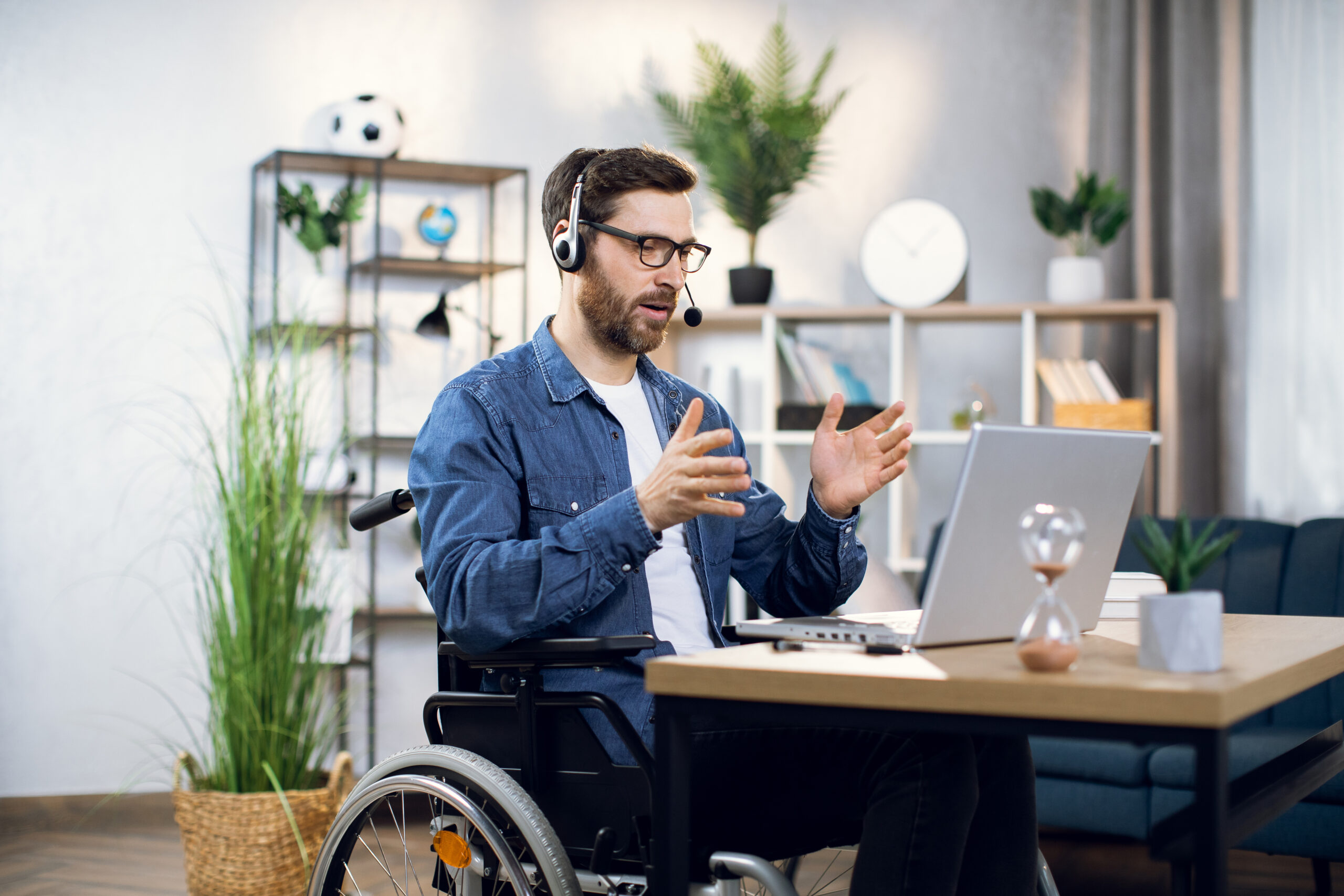 Bearded man in a wheelchair with eyeglasses and headset talking and gesturing during video conference on a laptop.